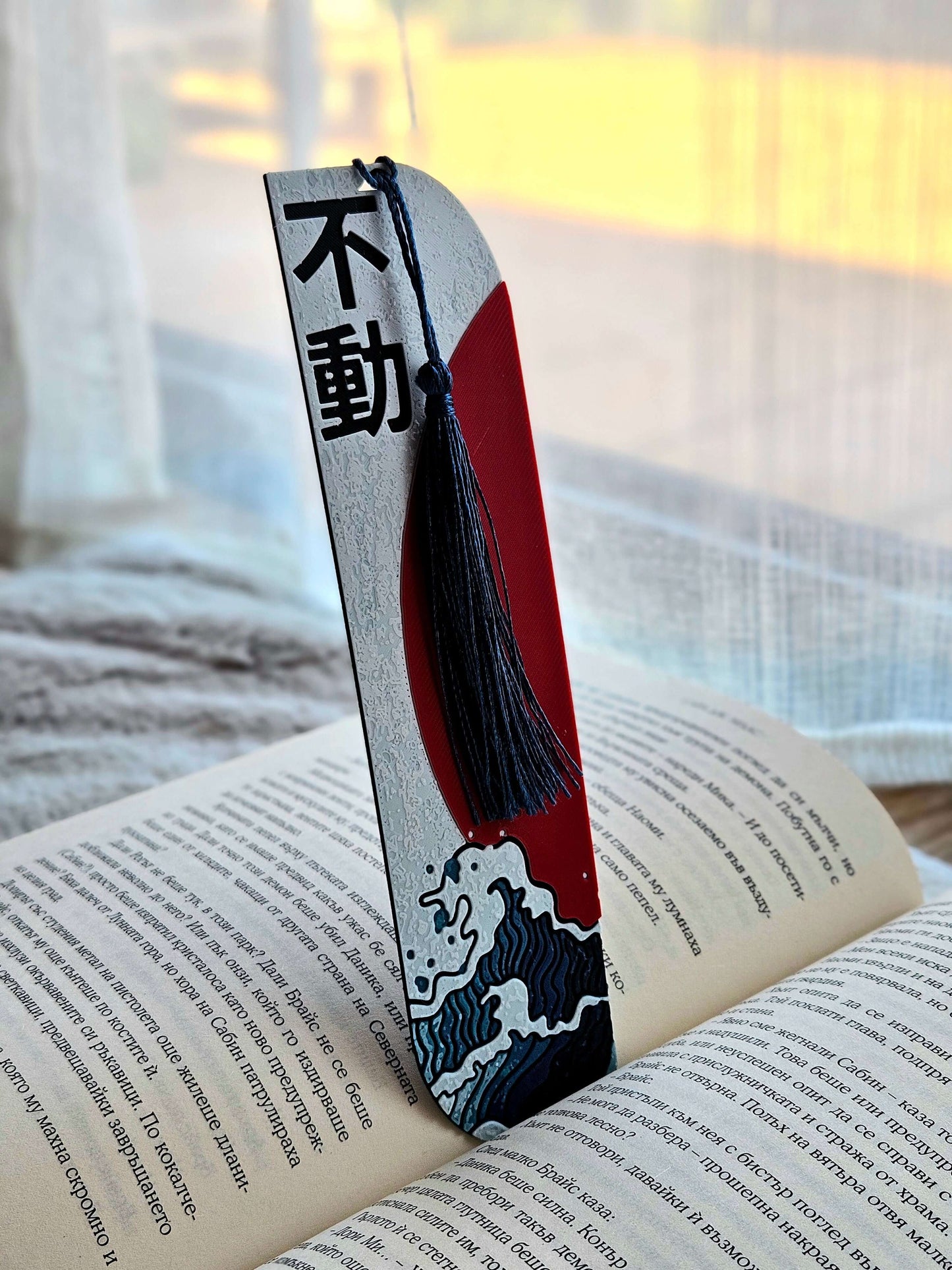 Semn de carte „Unshakable” (不動) cu design japonez, valuri și soare roșu, pe pagină deschisă.
